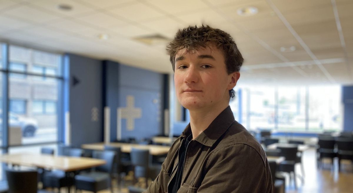 Young man with dark hair wearing brown jacket, smiling to camera, with college cafe in background